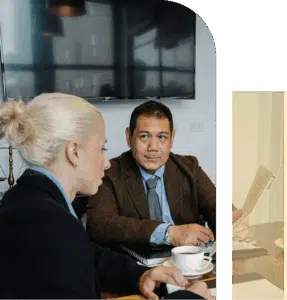 A blonde woman and an Asian man sit at a conference table with coffee, leaning in while discussing work in a shared meeting