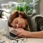 A woman rests her head on a desk beside an open laptop and glasses, eyes closed, showing mental fatigue after long work hours