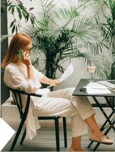 A woman works in a minimalist office or café, holding documents and a phone while concentrating on multiple responsibilities