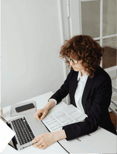 Curly-haired woman participates in a work meeting with laptop, phone, and organizer open, managing tasks in a busy setting