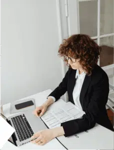 Curly-haired woman participates in a work meeting with laptop, phone, and organizer open, managing tasks in a busy setting