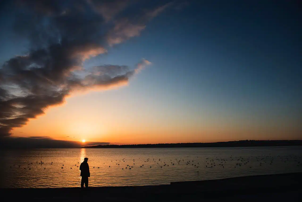 Person standing by calm water at sunset, reflecting emotional relief and clarity through stress therapy