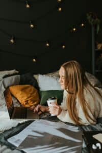 A woman working on her bed with papers spread around and a cup of coffee nearby, blending focus and fatigue