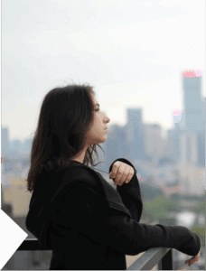 Woman standing on a balcony with New York City buildings behind her, contemplative stance facing the urban view
