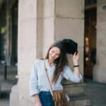 A woman standing beside a building pillar, removing her cap while looking down with a soft smile
