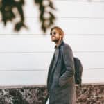 Man wearing a backpack standing before a white wall with leaves in the foreground, smiling in soft natural light