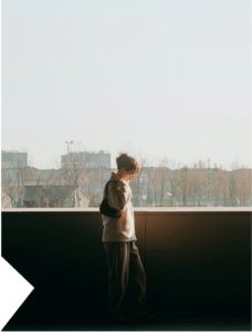 A woman standing quietly on a balcony with bright light behind her and shadow beneath, creating a reflective urban atmosphere
