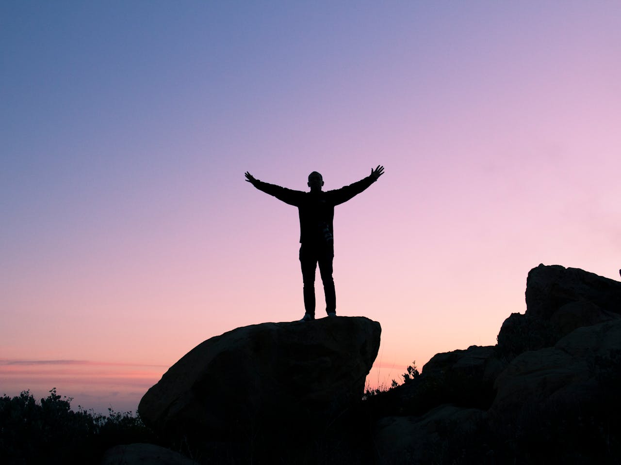 Silhouette standing on a rock at sunset with arms open, symbolizing resilience and relief from pressure to succeed