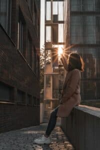 A woman seated on a balcony raising her hand toward the sun with city buildings behind her, creating a peaceful urban moment