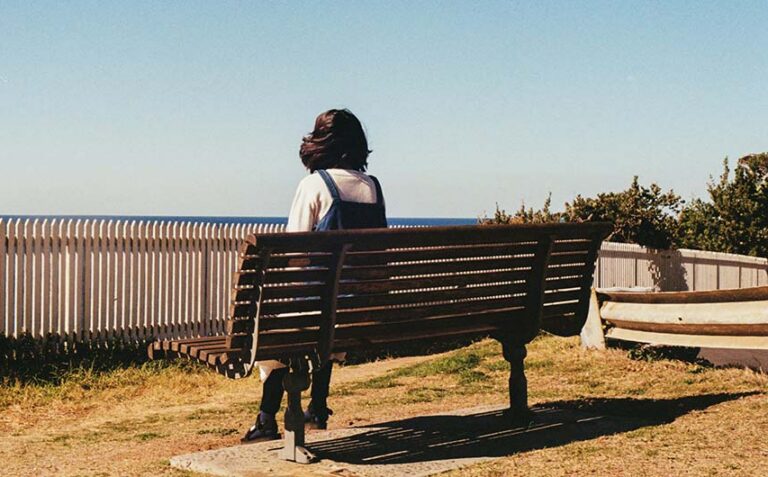 A person sits on a wooden park bench, facing away from the camera and looking out toward a clear blue horizon where the sky meets the ocean.