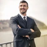 Confident male professional in a dark suit and tie standing outdoors with crossed arms, smiling.
