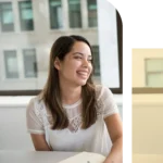 Smiling young professional woman with long dark hair wearing a white lace top sitting at an office desk.