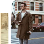 Young man with glasses wearing a brown coat holding a DSLR camera and phone while standing on an urban crosswalk.
