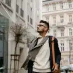 A stylish man wearing autumn streetwear layers and a leather messenger bag walking through a European city on his daily commute.