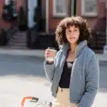 A young woman with curly brown hair wearing a blue jacket stands next to a bicycle on a city street, holding a takeaway coffee cup